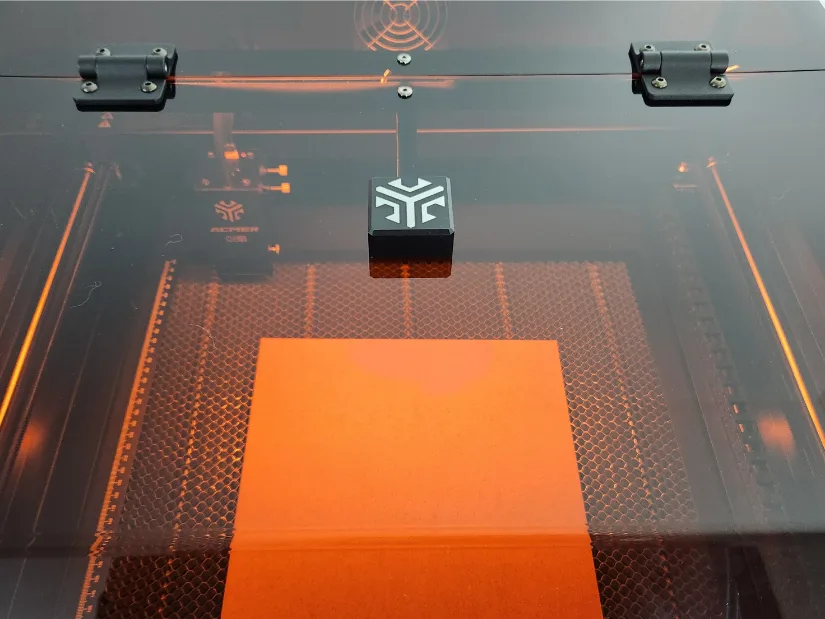 Top-down view through the closed amber lid of the Acmer P3 laser engraver showing the laser module, work area with honeycomb bed, and an orange test sheet positioned for engraving