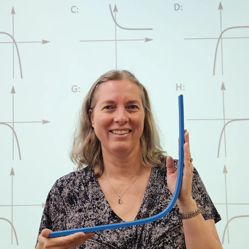 Math teacher holding a large 3D printed exponential curve model in front of coordinate graphs during a classroom lesson